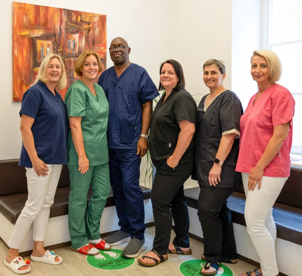 Sechs medizinische Fachkräfte in unterschiedlichen Farben der Arbeitskleidung stehen lächelnd vor einem farbenfrohen Gemälde, auf dem Boden sind Markierungen sichtbar.